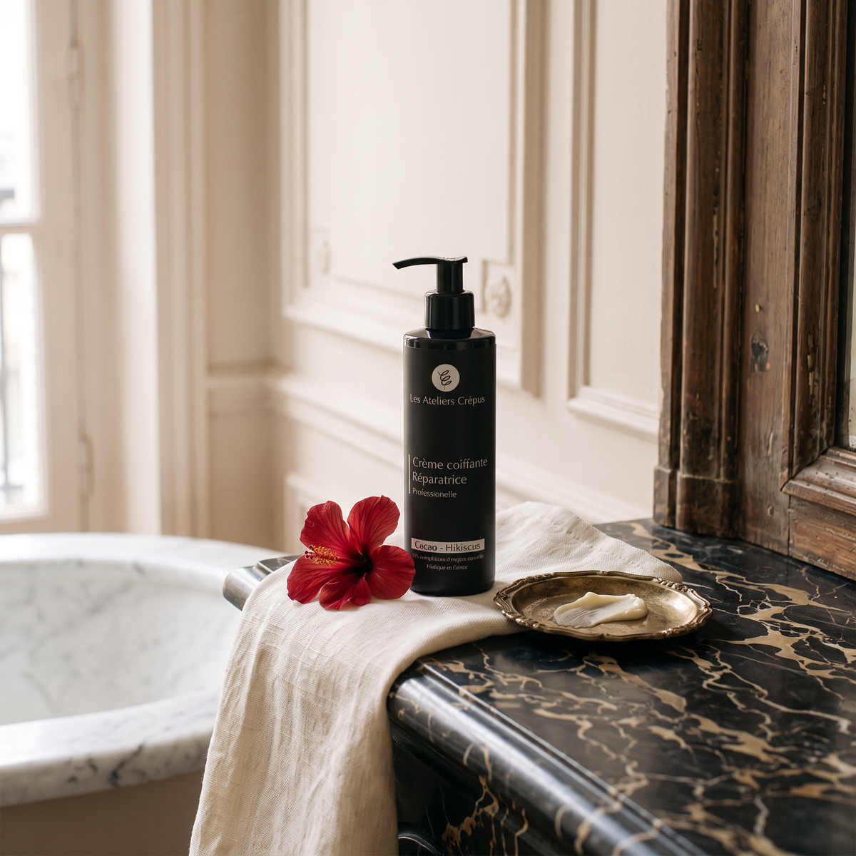 La Crème Coiffante posée sur une commode en marbre noir, avec une fleur d'hibiscus, devant une baignoire