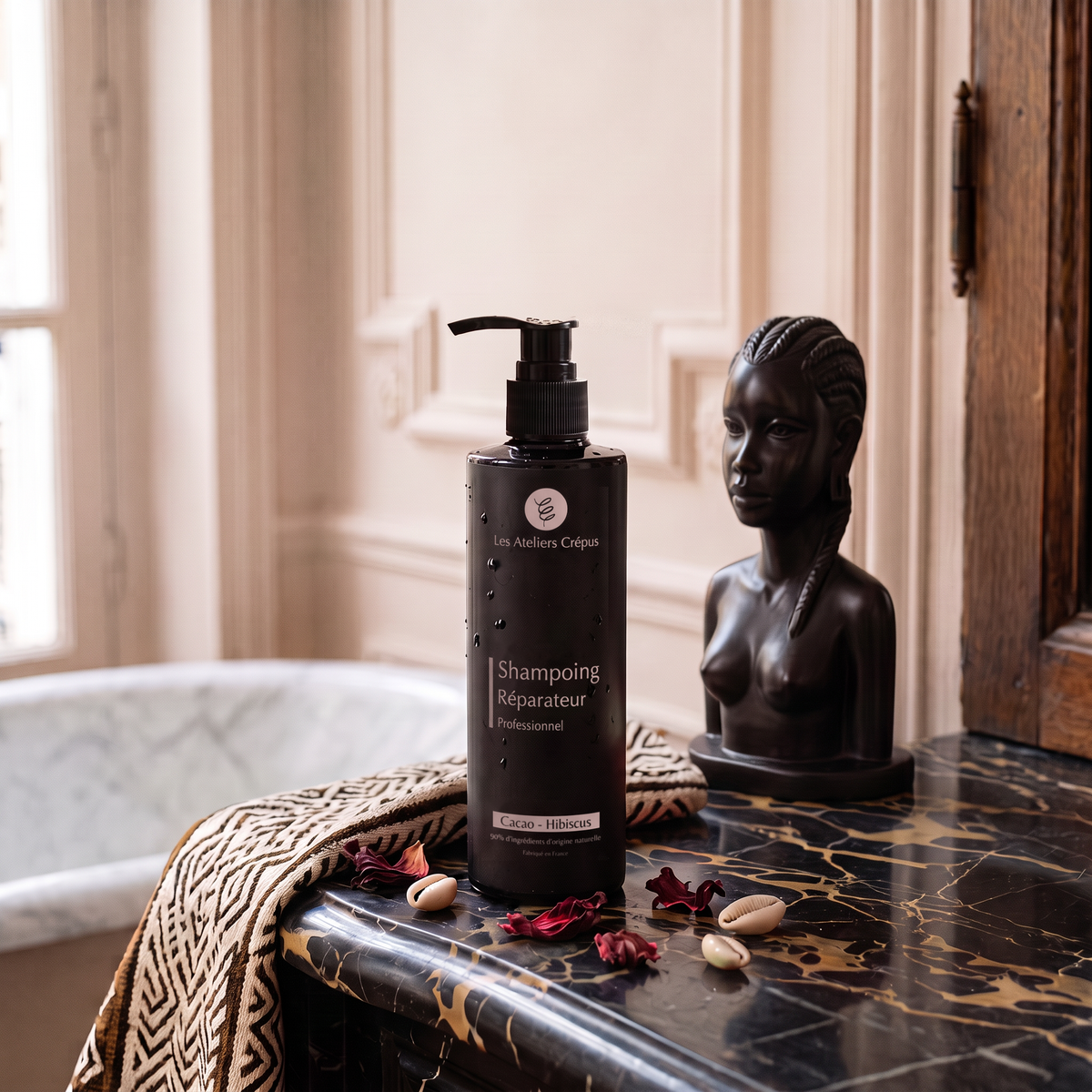 Le Shampoing Réparateur Cacao Hibiscus posé sur une commode en marbre noir, devant une baignoire, avec une statuette et un tissu imprimé