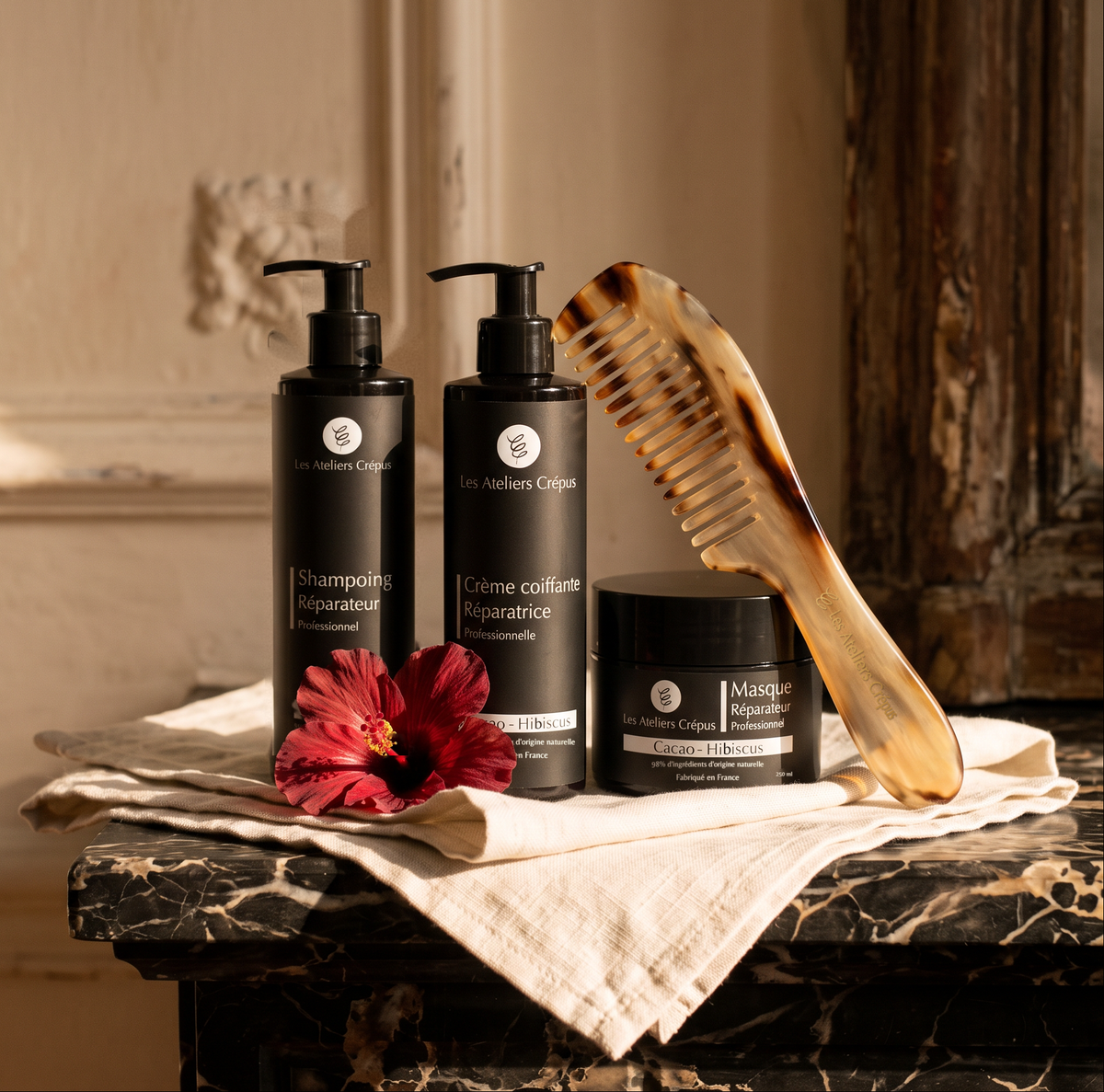 Rituel Texture® et son Peigne : Le Shampoing, La Crème Coiffante, Le Masque Réparateur et le Peigne de Man Dol’ sur une commode en marbre, avec des fleurs d'hibiscus