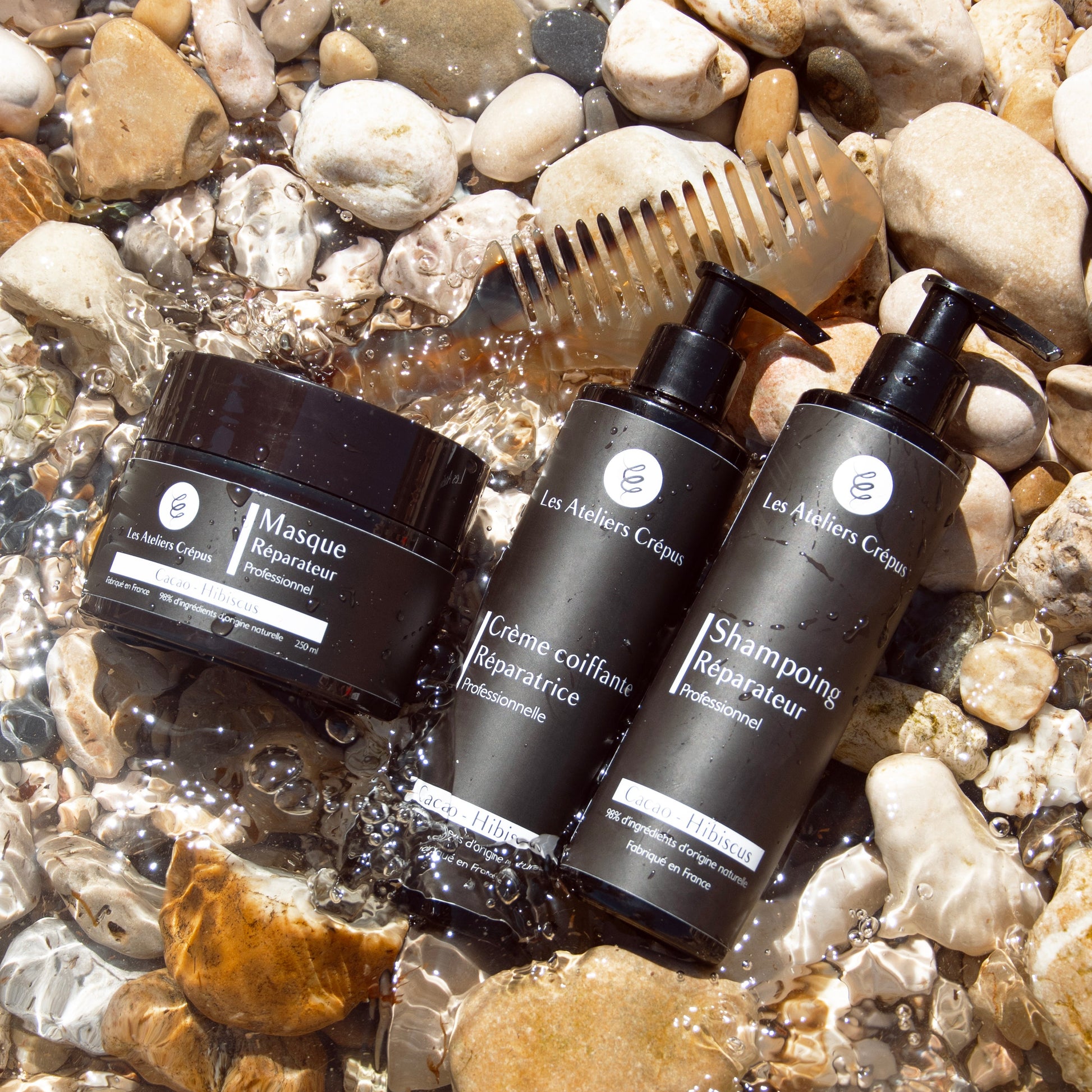 Gamme cheveux Les Ateleirs CRépus masque réparateur, peigne en corne chamspoing hydratant et crème coiffante posés dans un ruisseau