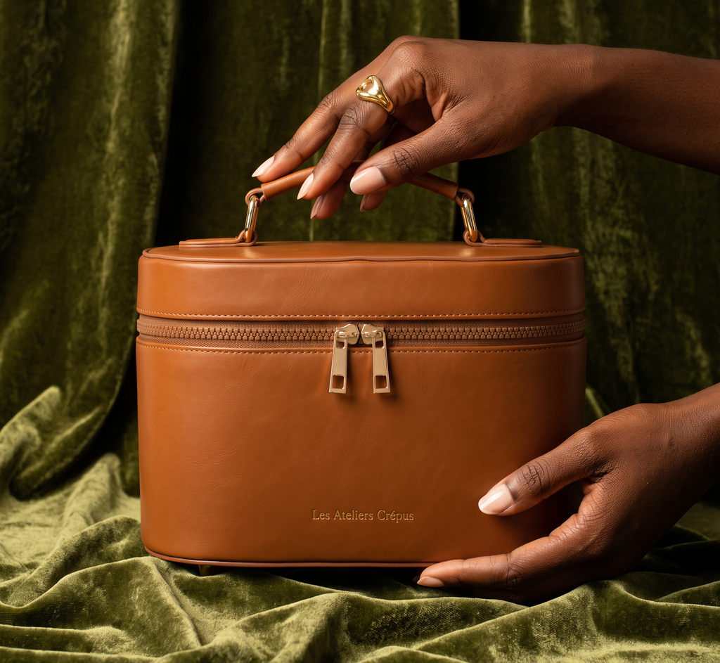 Main sur la poignée du Vanity de Man Dol’ en cuir camel, posé sur un tissu vert